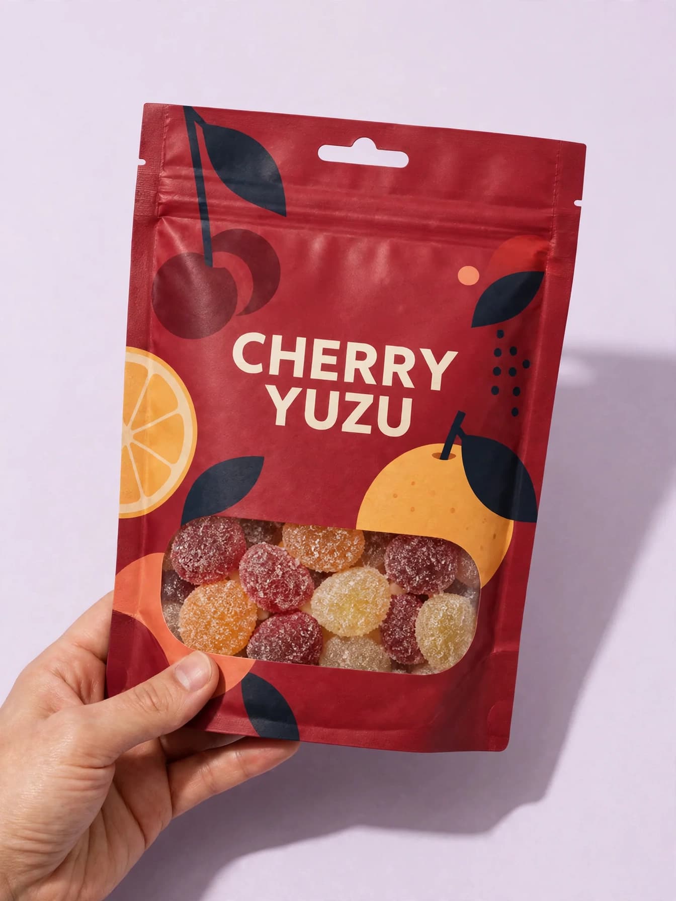 Hand holding a cherry and yuzu gummy pouch against a pale lilac studio background